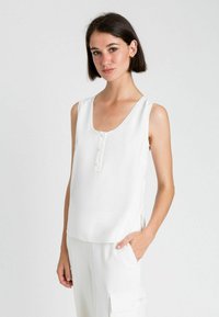 Top bianco senza maniche con scollatura a barca, dotato di bottoni sul davanti e di uno spacco laterale per facilitare il movimento. Tessuto dalla texture liscia.