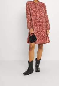 Šaty s potiskem zvířecí kůže v červené a krémové barvě, s dlouhými rukávy, výstřihem ve tvaru klíče a rozšířeným lemem. K šatům jsou obuty černé kovbojské boty a malá černá kabelka.