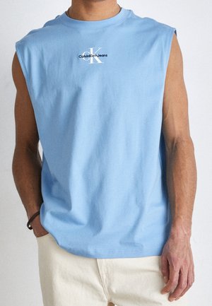 Mann trägt ärmelloses hellblaues Calvin Klein Jeans Hemd und cremefarbene Hose mit einer Hand in der Tasche vor weißem Hintergrund.