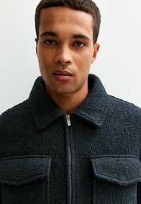 Czarna, strukturalna kurtka z zamkiem z przodu, spiczastym kołnierzem i dwiema kieszeniami na piersi. Materiał wygląda na gruby i ciepły, z miękkim wykończeniem.