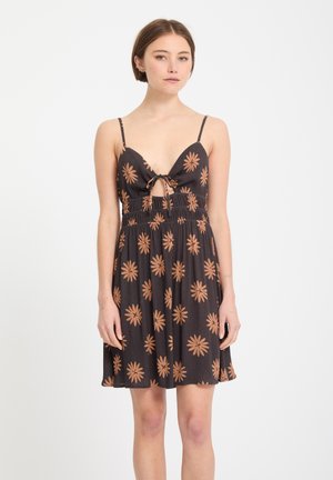 Jeune femme portant une robe noire sans manches avec un motif floral marron et un détail noué à l'avant, debout devant un fond blanc.