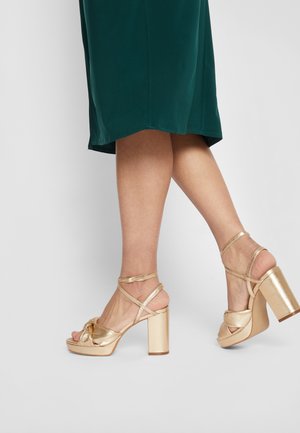 Sandalias doradas de tacón cuadrado; cuentan con tiras cruzadas, un detalle de nudo en la parte delantera y un acabado metálico brillante.