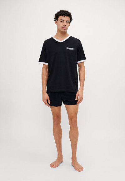 T-shirt preta com bordas brancas, decote em V e logotipo frontal. Usada com calções escuros. O tecido parece suave e leve.