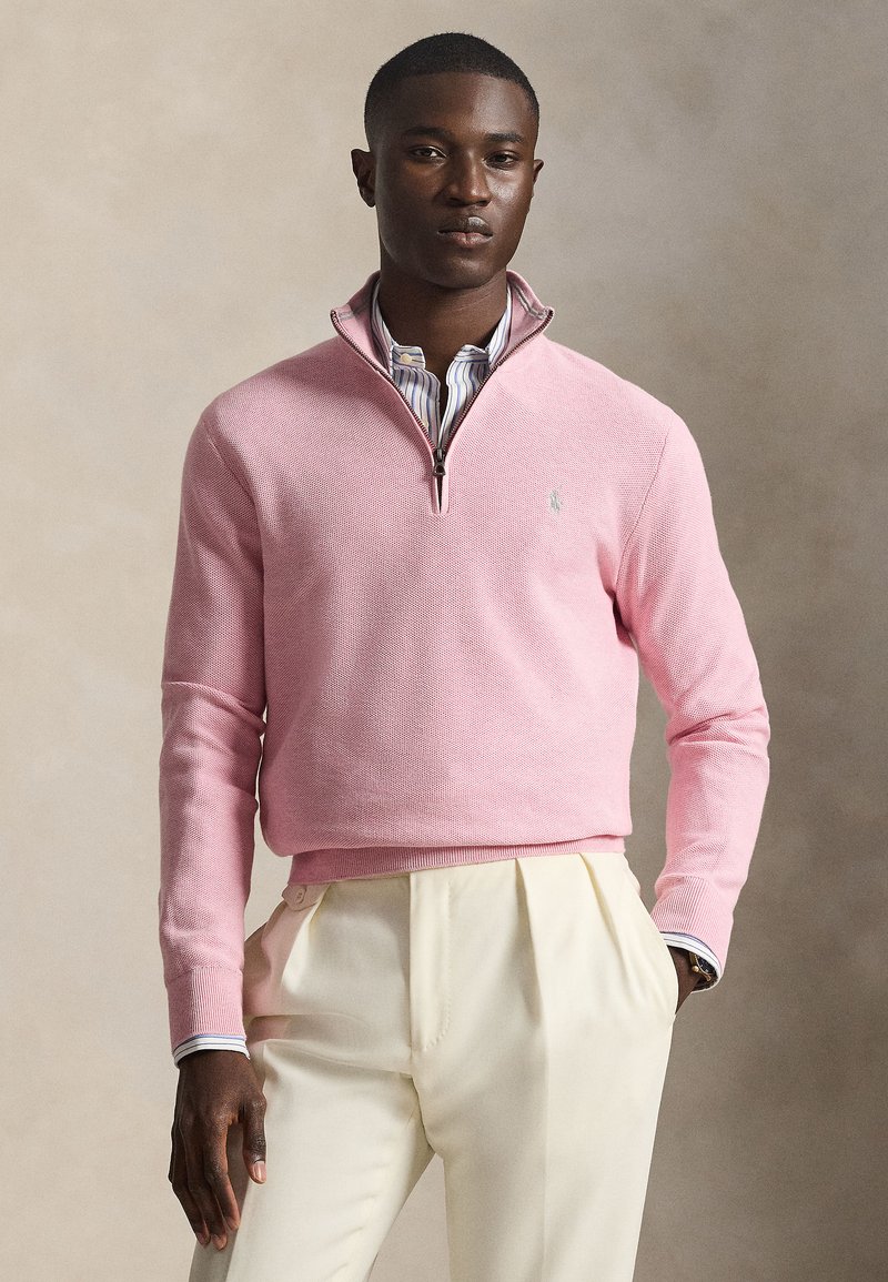 Ανοιχτό ροζ πουλόβερ με φερμουάρ σ το λαιμό, πλεκτή υφή, μανσέτες με ραβδώσεις, φορεμένο πάνω από ριγέ πουκάμισο, συνδυασμένο με κρεμ παντελόνια.