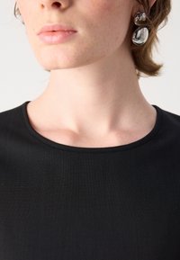 Black, short-sleeve top with a smooth texture and round neckline, paired with shiny, silver drop earrings. Close-up of neck and shoulders.