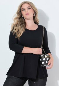 Schwarzes Langarmoberteil mit rundem Halsausschnitt. Das Modell hält eine schwarze Handtasche mit Punkten und goldenen Details, kombiniert mit verzierten dunklen Jeans.