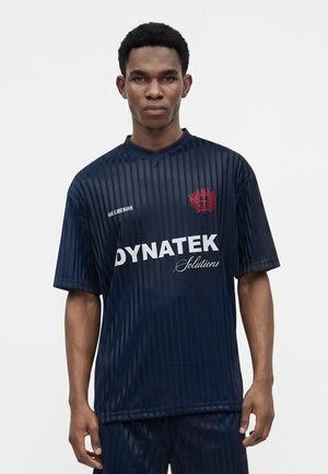 Jeune homme portant un maillot de sport rayé bleu foncé avec un emblème rouge et le logo "DYNATEK Solutions", debout sur un fond blanc uni.