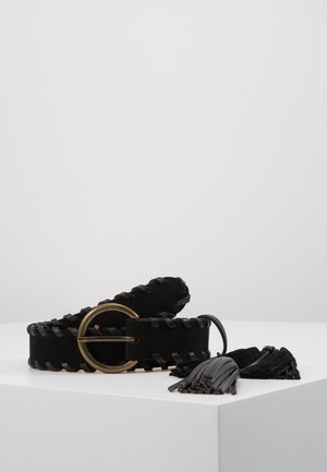 Ceinture tissée noire avec boucle ronde en laiton et extrémités à pampilles, présentée enroulée sur une surface blanche avec un fond gris clair.