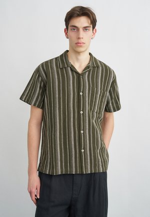 Młody mężczyzna o krótkich brązowych włosach, ubrany w zieloną i białą koszulę w pionowe paski z krótkim rękawem i ciemne spodnie.