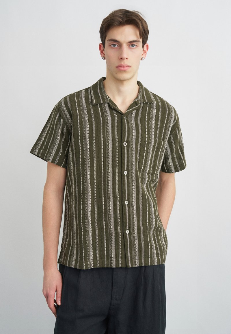 Νεαρός άνδρας με κοντά καστανά μαλλιά, φορώντας πράσινο και λευκό πουκάμισο με κάθετες ρίγες και κοντά μανίκια και σκούρο παντελόνι.