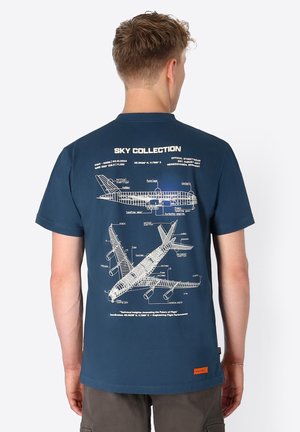 Man die een donkerblauw T-shirt draagt met technische vliegtuigdiagrammen en labels op de rug, staand met zijn rug naar de camera.