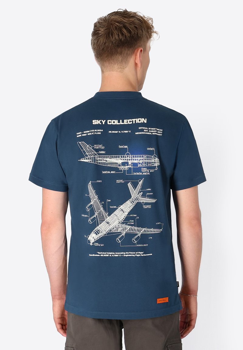 Mann trägt ein dunkelblaues T-Shirt mit technischen Flugzeugdiagrammen und Beschriftungen auf dem Rücken, steht mit dem Rücken zur Kamera.