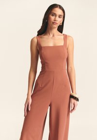 Roestkleurige jumpsuit met een vierkante halslijn, brede schouderbanden en taps toelopende pijpen. Gemaakt van een gladde, gestructureerde stof. Voorzien van zijzakken.