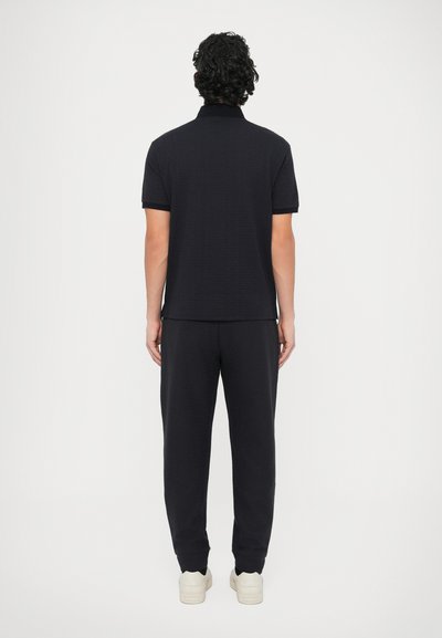 Μαύρο πόλο πουκάμισο και παντελόνι σε ταιριαστό υφασμα με υφή. Το σύνολο ολοκληρώνεται με ανοιχτόχρωμα αθλητικά παπούτσια.