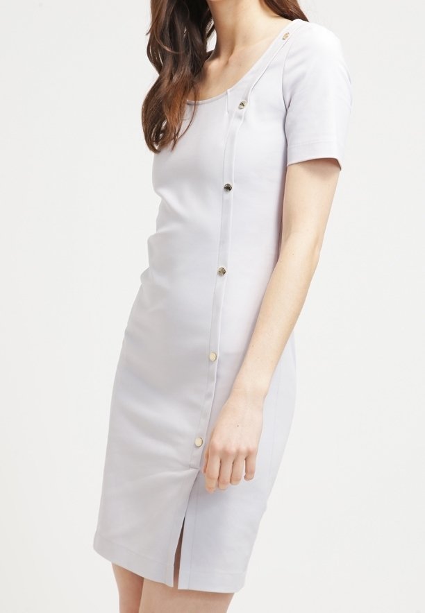 Femme portant une robe ajustée gris clair à manches courtes avec des boutons dorés disposés en diagonale de l'épaule à l'ourlet.