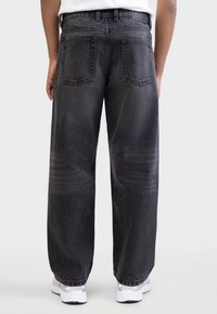 Blugi negri din denim cu picioare largi și o textură estompată, având două buzunare în spate și o buclă pentru curea. Purtați cu adidași albi.