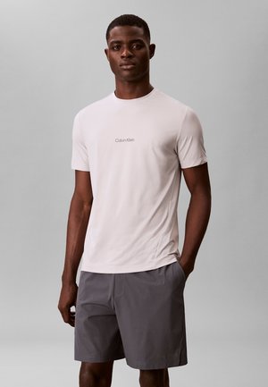 Ung mand står med hænderne i lommerne iført en lys beige Calvin Klein T-shirt og mørkegrå shorts mod en ensfarvet baggrund.