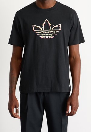 Mann trägt ein schwarzes T-Shirt mit kurzen Ärmeln und einem neonpinken und gelben stilisierten Flammenlogo in der Mitte der Brust.