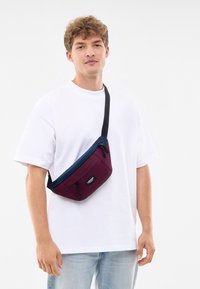 Bolso bandolera en rojo y azul marino con forma rectangular, compartimentos con cremallera y una correa negra ajustable. El material parece duradero y texturizado.
