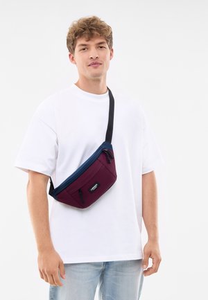 Röd och marinblå crossbody-väska med rektangulär form, dragkedjfack och en justerbar svart rem. Materialet verkar slitstarkt och texturerat.