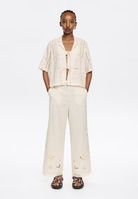 Beige cropped top met striksluiting en ingewikkelde borduursels, gecombineerd met beige wijde broek met bloemen details. Draagt bij kleurrijke sandalen.