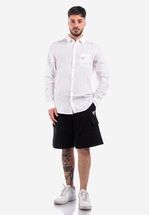 Camicia bianca di lino con bottoni e tasca, abbinata a pantaloni cargo neri con tasche laterali. Il modello indossa sneakers bianche.