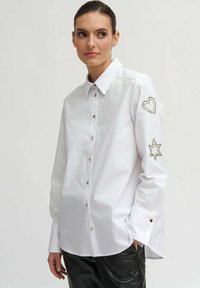 Biała koszula zapinana na guziki z klasycznym kołnierzykiem, długimi rękawami ozdobionymi wzorami serc i gwiazd z ćwiekami oraz brązowymi guzikami z przodu.
