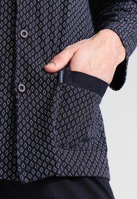 Marineblauw shirt met een wit en rood geometrisch diamantpatroon, met knoopsluiting aan de voorkant en een borstzak. Gemaakt van zachte stof met een gladde textuur.