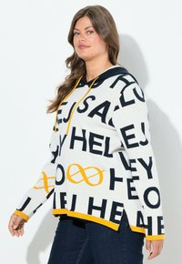 Femme avec de longs cheveux bruns portant un sweat à capuche blanc avec des lettres noires et des symboles d'infini jaunes, associé à un jean bleu foncé.