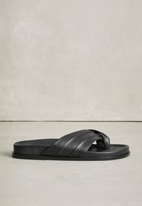 Sandale slide en cuir noir avec larges brides croisées et semelle plate sur un sol lisse de couleur claire, devant un mur uni.