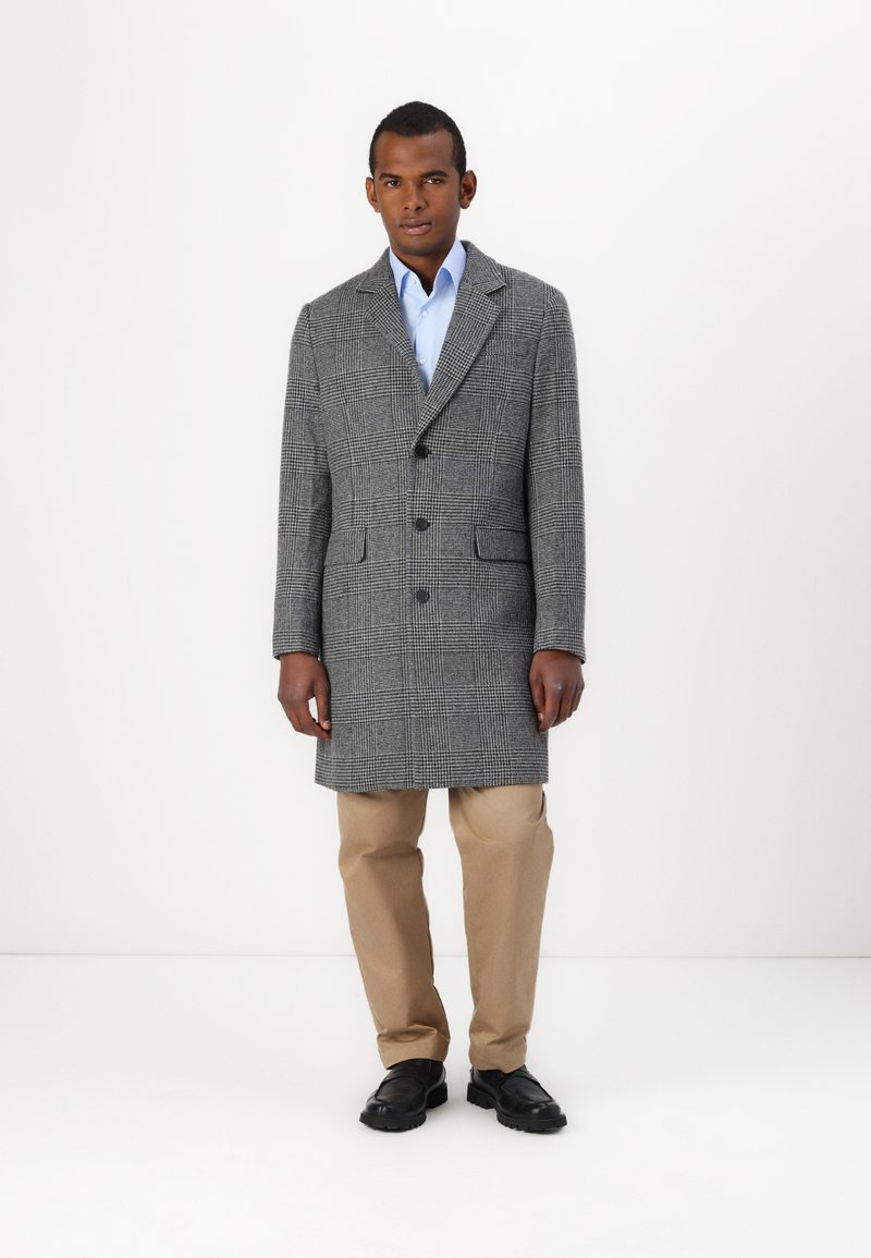 Gray houndstooth overcoat with notch lapels, two buttons, and side pockets; worn over a light blue shirt, paired with beige trousers and black shoes.
