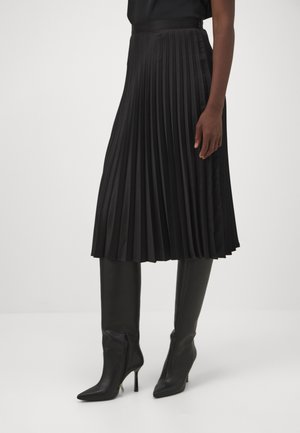 Frau trägt einen schwarzen plissierten Midirock und schwarze kniehohe Spitzabsatzstiefel und steht vor einem weißen Hintergrund.