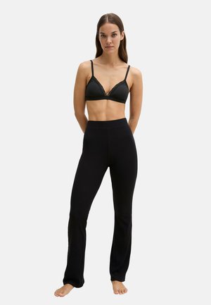 Femme debout pieds nus, portant un bralette noir avec des bordures en dentelle et un pantalon noir taille haute sur un fond clair uni.