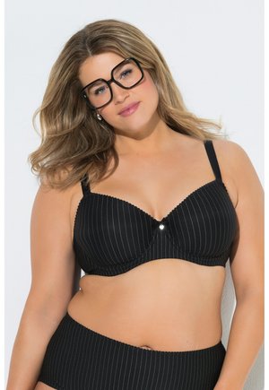 Woman with wavy hair wearing large black glasses, a black pinstripe bra, and matching high-waisted underwear against a white background.