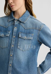 Camicia in denim azzurro chiaro con bottoni a pressione di perla, due tasche frontali e logo ricamato sulla tasca destra, con un design di colletto classico.
