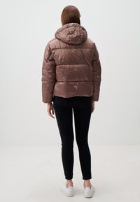 Braune Pufferjacke mit Kapuze, die ein gestepptes Design und florale Akzente aufweist. Modell trägt schwarze Skinny Jeans und weiße Sneakers.