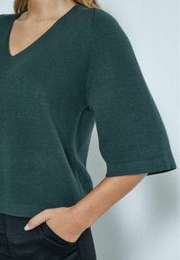 Top verde lavorato a maglia con scollatura a V e maniche corte e larghe. Ha una vestibilità comoda e una texture morbida, abbinato a pantaloni neri.