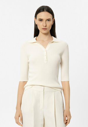 Frau mit glatten dunklen Haaren, die ein cremefarbenes, kurzärmeliges Rippen-Poloshirt und passende, hoch taillierte, plissierte Hosen trägt, frontal stehend.