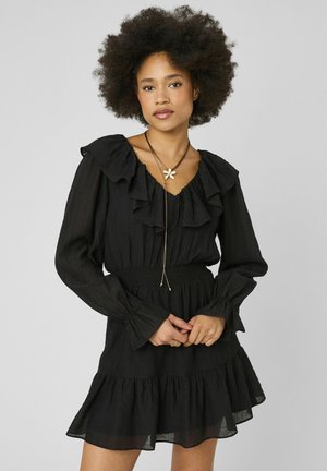 Femme avec coiffure afro portant une robe noire à manches longues à volants et un long collier avec un pendentif fleur, debout les mains jointes.