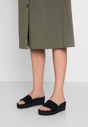 Czarne sandały na platformie z teksturowanymi paskami, odkrytymi palcami i grubą podeszwą. Noszone z khaki spódnicą sięgającą kolan.