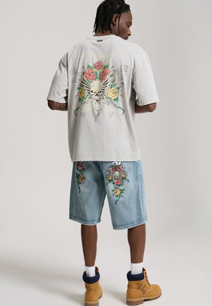 Mężczyzna z zaplecionymi włosami, ubrany w beżową koszulkę z nadrukiem czaszki i róż, pasujące kwiatowe szorty jeansowe, białe skarpetki i jasnobrązowe buty, stoi tyłem.