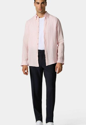 Chemise en lin rayée rose clair à manches longues et à boutons, portée sur un T-shirt blanc et associée à un pantalon sombre et des baskets blanches.