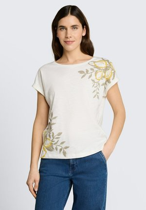 Vrouw draagt een wit kortemouwen top met gele en grijze bloemenprints, gecombineerd met blauwe jeans, staand met één hand in de zak.
