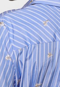 Ljusblå randig skjorta med broderade bi-accenter och paljetter. Tyget verkar slätt och lättviktigt med en klassisk krage.