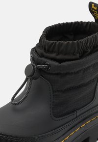 Juodas vandeniui atsparus batas su tekstūruota nailono viršūne, suspausta elastinga apykakle ir storu guminiu padu su geltonais siūlės akcentais.