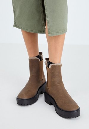 Bruine leren enkellaarzen met zwarte robuuste zolen en een voering van bont, gedragen met een groene rok en zwarte sokken op een witte vloer.