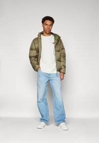 Chaqueta aislante en verde oliva con capucha, camiseta blanca de cuello redondo y jeans de corte amplio en azul claro. Las zapatillas blancas completan el conjunto.