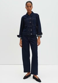 Giacca in denim scuro e pantaloni abbinati con vestibilità classica, dotati di chiusura frontale con bottoni, due tasche sul petto e polsini arrotolati. Scarpe nere slip-on con borchie.