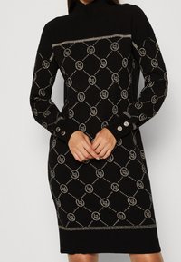 Kvinna i svart stickad klänning med silver geometriskt och cirkulärt mönster, knoppar på ärmarna och hög hals, står med händerna ihopknutna.