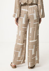 Pantaloni a gamba larga in un tessuto bianco leggero con un motivo geometrico marrone. Vita elasticizzata e vestibilità comoda. Sandali piatti visibili.